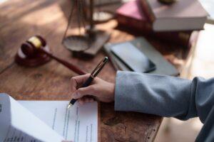 Lawyer's hand holding a pen and signing a legal contract, agreement, or will, with a gavel, scales of justice, and law books in the background, representing legal process and advice