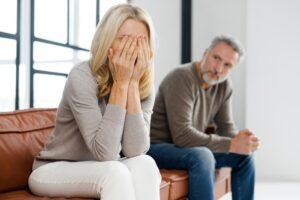 Upset middle-aged couple is arguing at home, frustrated blonde woman covered face with palms and crying, sadness grey-haired mature man on background.